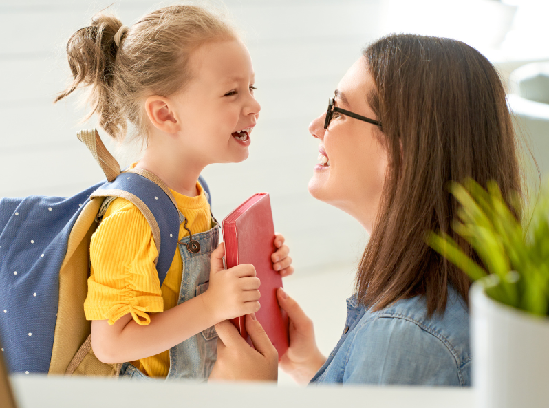 Uśmiechnięta dziewczynka stoi z zeszytem w rękach, a przy niej kuca mama.