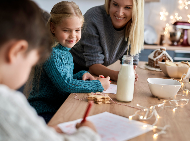 Dzieci piszą list do Świętego Mikołaja, obok stoi ich mama, na stole ozdoby świąteczne